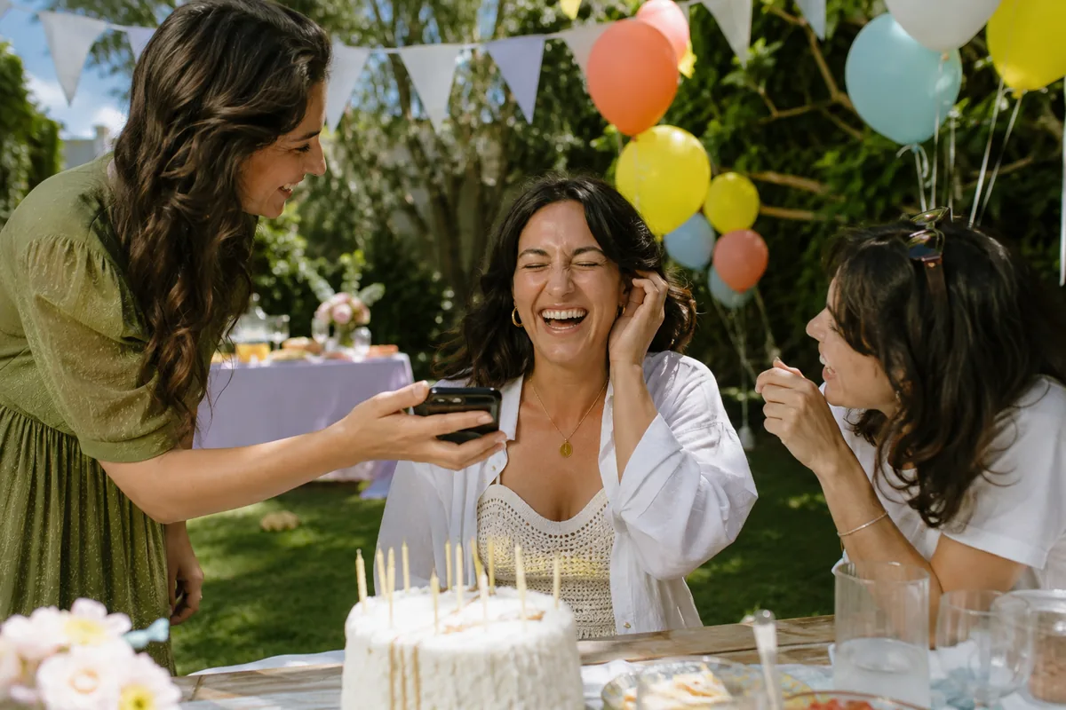 A woman laughing with her eyes closed at a garden birthday party as two friends hold a phone up to her, coral, blue and yellow balloons and white bunting behind, a cake with unlit candles on the table.