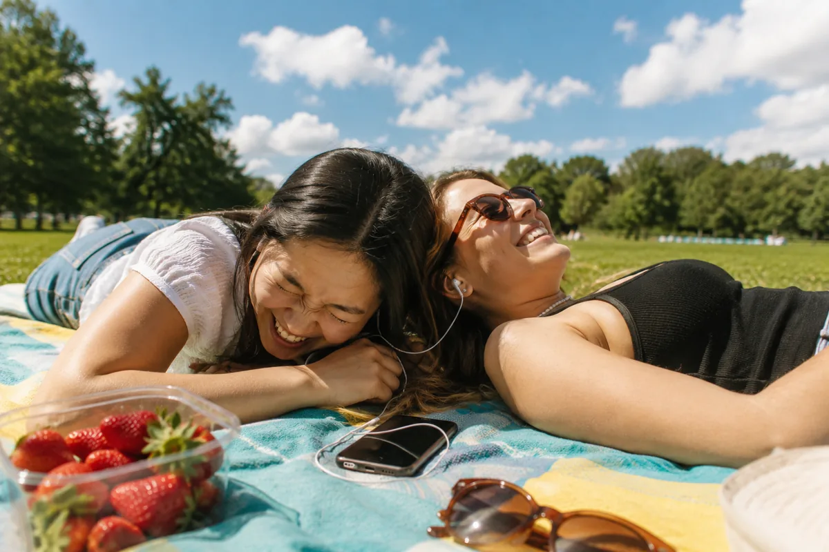 Two friends lying head to head on a turquoise picnic blanket in a sunny park sharing one pair of earphones, one laughing into the blanket, the other grinning up at the sky, strawberries and sunglasses beside them.