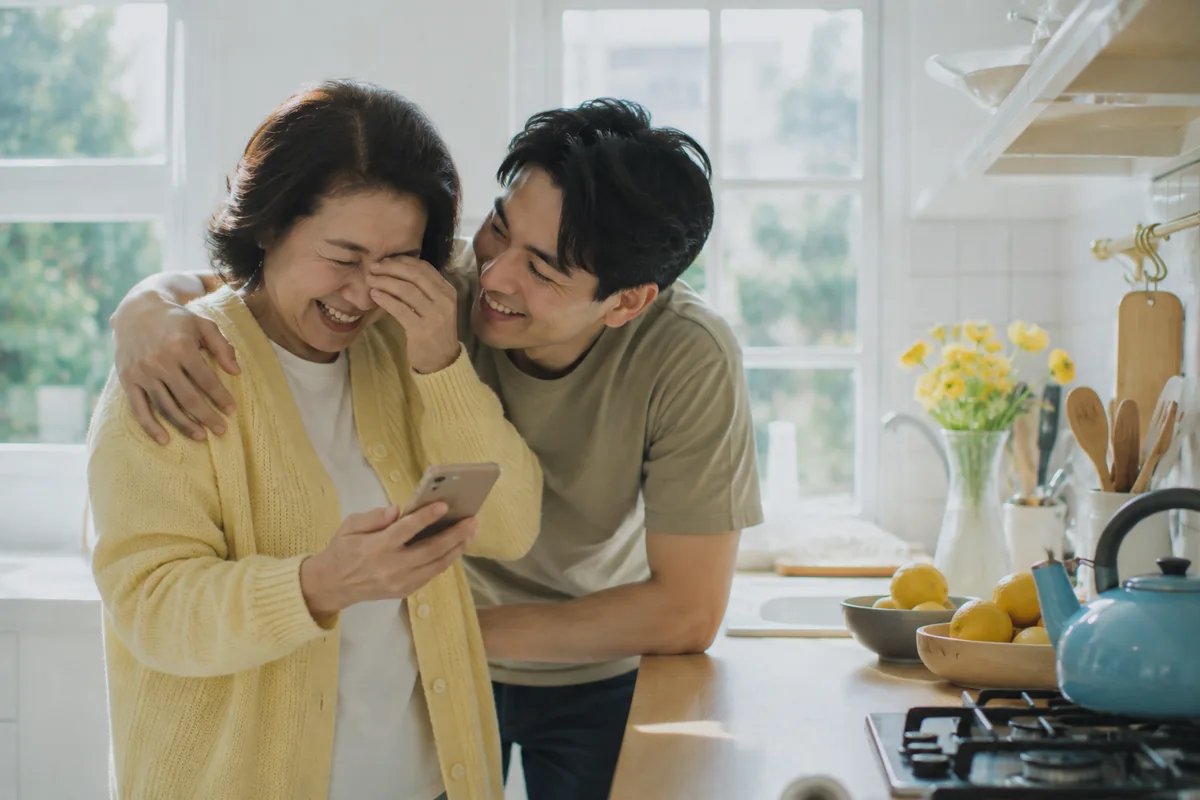 A mother in a yellow cardigan in a bright white kitchen wiping her eyes and laughing at the phone in her hand, her grown son hugging her from the side, yellow flowers and a blue kettle on the bench.
